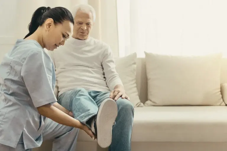 A home health aide helps an older man with his shoes in his living room
